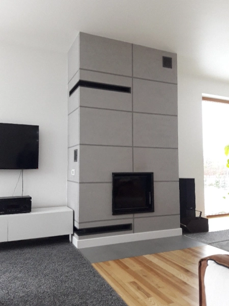 Nowoczesny kominek zabudowany w gładkiej, szarej konstrukcji z geometrycznym podziałem płyt. Wkład kominkowy umieszczony centralnie, a całość stanowi elegancki element salonu o minimalistycznym wystroju.